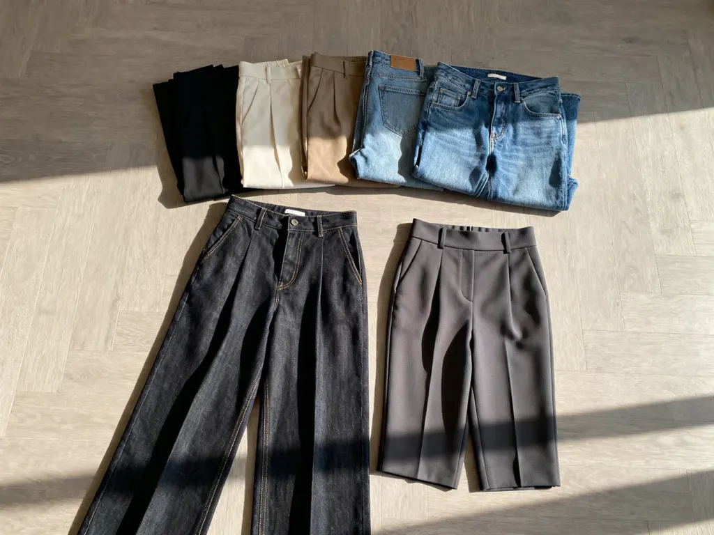 Różne modele spodni i jeansów z wysokim stanem ułożone na drewnianym podłożu, prezentujące propozycje dla różnych sylwetek.